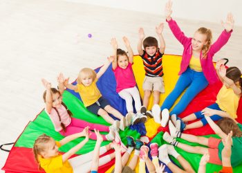 Portrait of young nursery teacher playing circle games with preschool children, sitting on rainbow parachute in gym